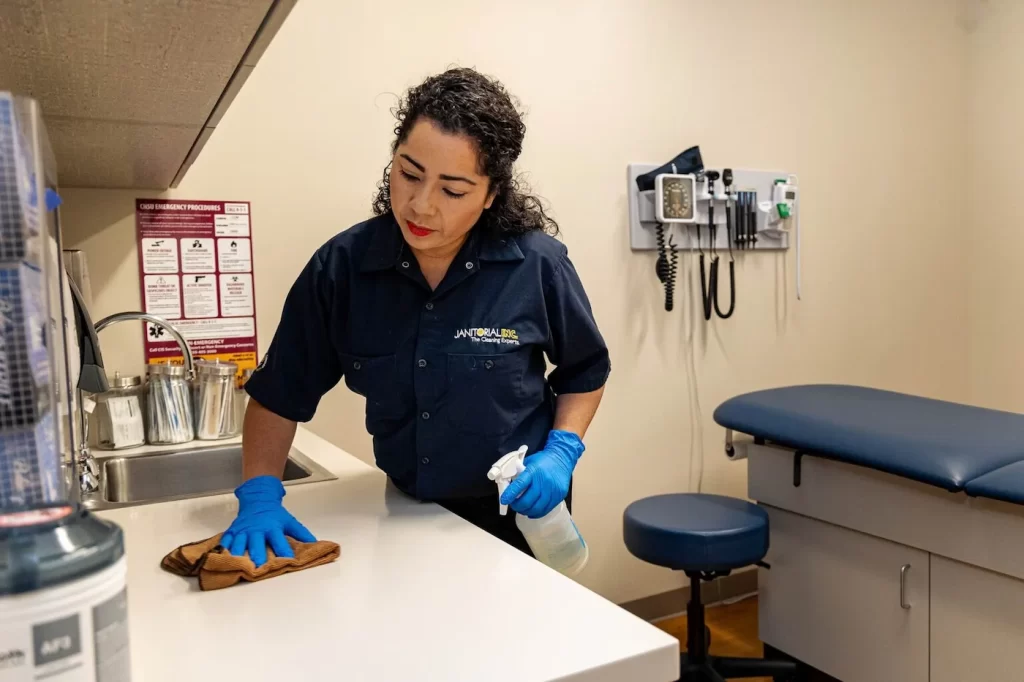 Janitorial woman cleaning surface of a table in a medical room.